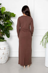 A woman with long dark hair faces away, wearing the Modest Goss Maya Tie-up Maxi Dress in cocoa brown. Indoors, she stands between two large potted plants against a white wall, showcasing modest fashion.