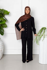 A woman smiles indoors wearing the Jasmine 2 Piece Set (Black) by Modest Goss and a brown hijab. She stands near green potted plants, a white textured pillar with a trailing plant, and white walls with decorative molding.