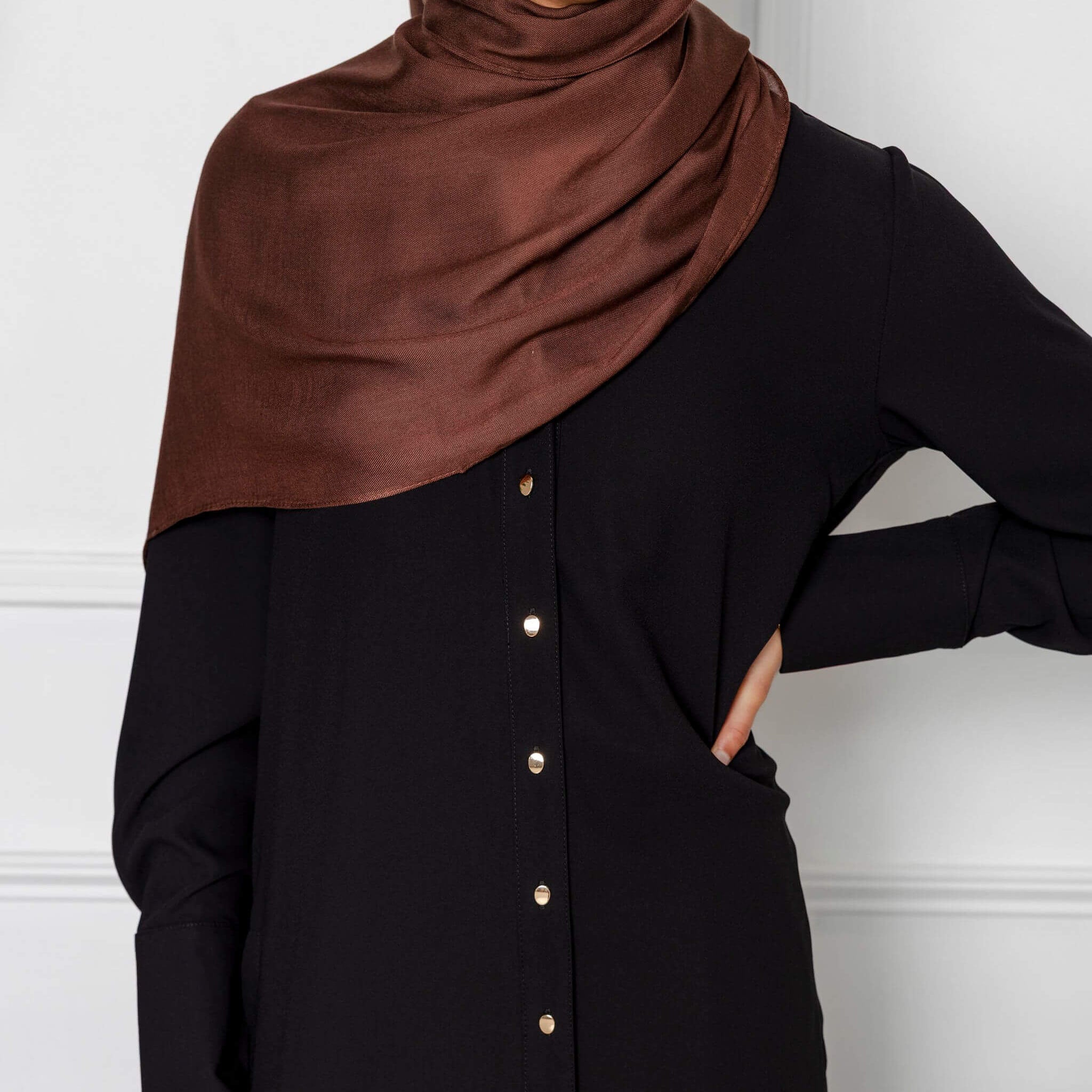 Wearing a dark brown satin hijab and the Modest Goss Jasmine 2 Piece Set in black with gold buttons, a person poses against a white wall with one hand on their hip, presenting a chic and modest look.