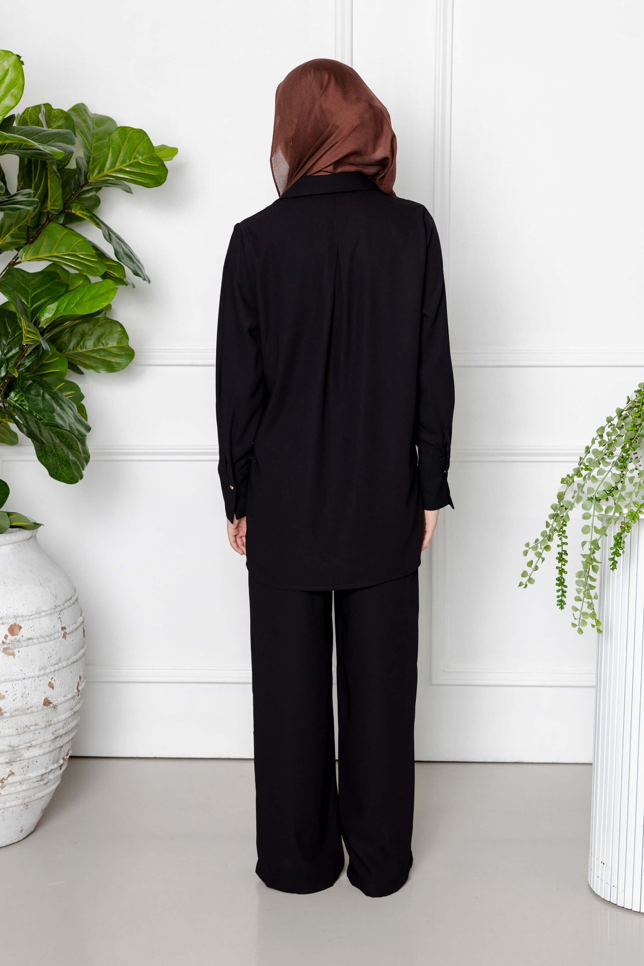 A person in the Modest Goss Jasmine 2 Piece Set (Black) and a brown headscarf stands with their back to the camera, positioned between two potted plants against a white paneled wall.