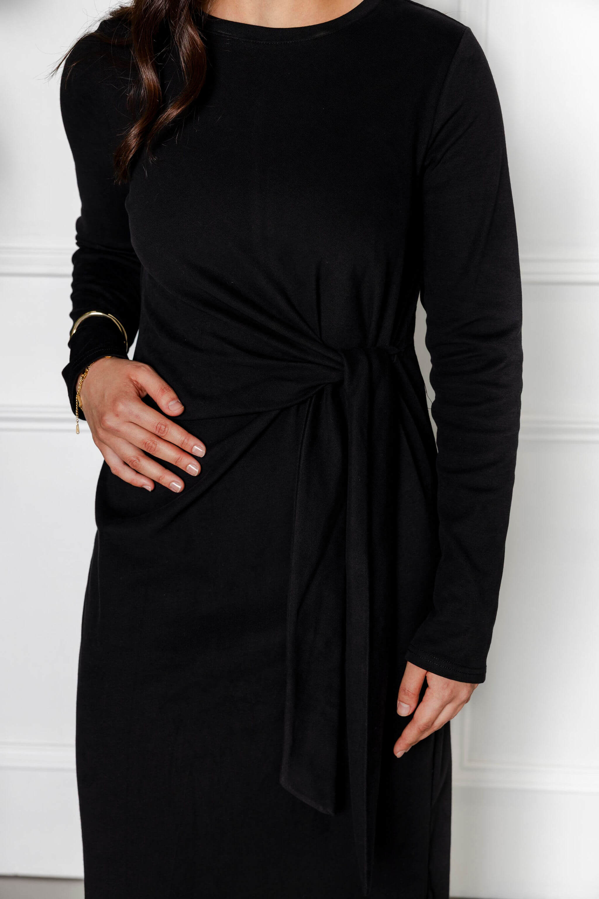 A woman models the Modest Goss Maya Tie-up Maxi Dress—a long-sleeved black dress with a waist knot—standing against a white background, her left hand on her hip and dark hair over one shoulder, showcasing contemporary modest style.