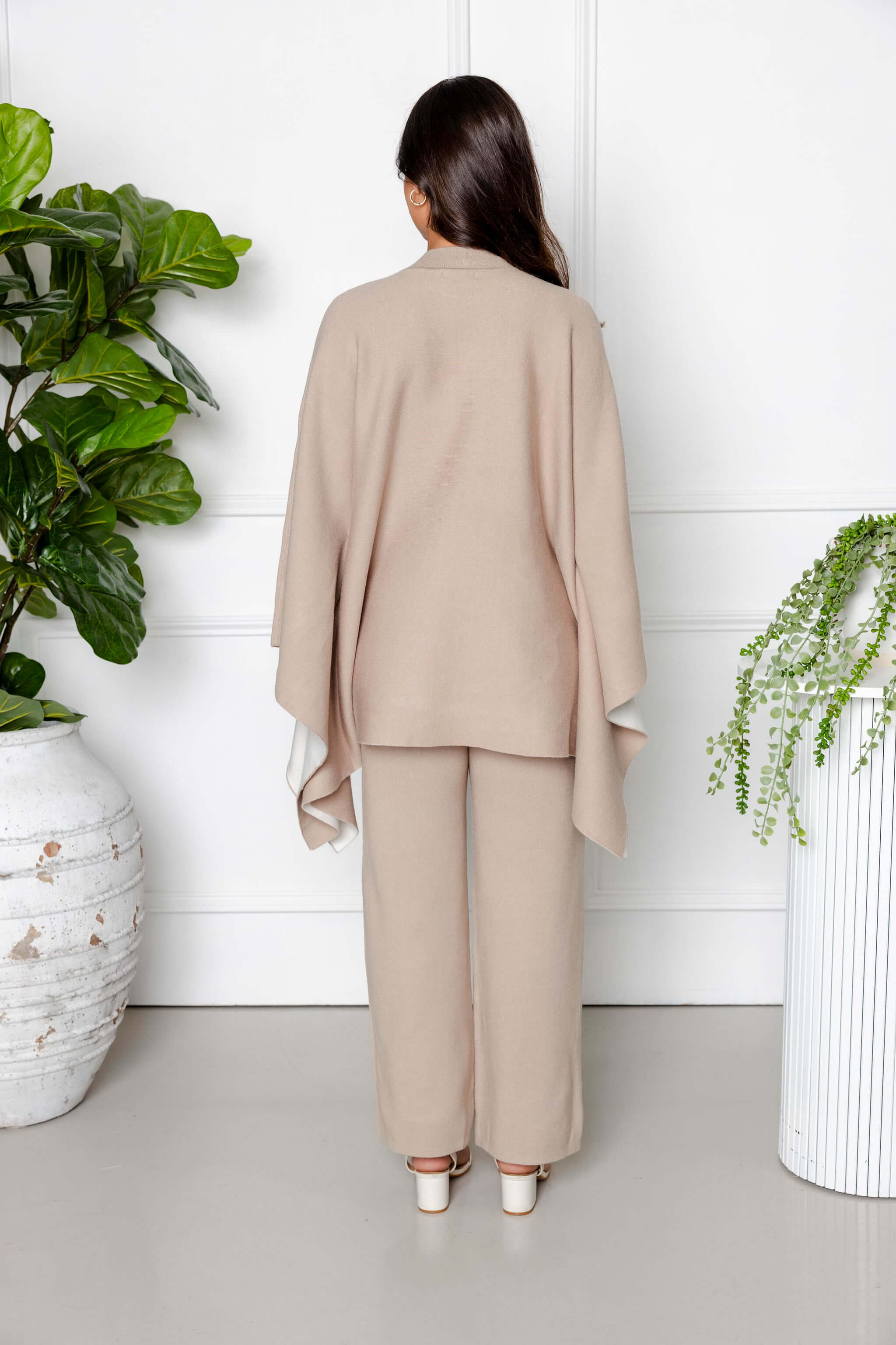 A woman with long dark hair stands indoors, facing away, wearing the Modest Goss Amara Knit Poncho 2 Piece Set in beige, featuring wide sleeves and pants. She is surrounded by green potted plants against a white wall.