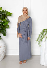 A woman in a beige hijab and the Maya Tie-up Maxi Dress by Modest Goss stands indoors between two large potted plants, smiling at the camera against a white wall, showcasing elegant modest fashion.