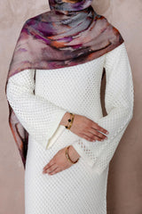 A person wears the Noor Knit Maxi Dress in white by Modest Goss with a purple and orange patterned scarf, standing against a beige background, arms crossed and gold bracelets on their wrists. Their face is not visible.