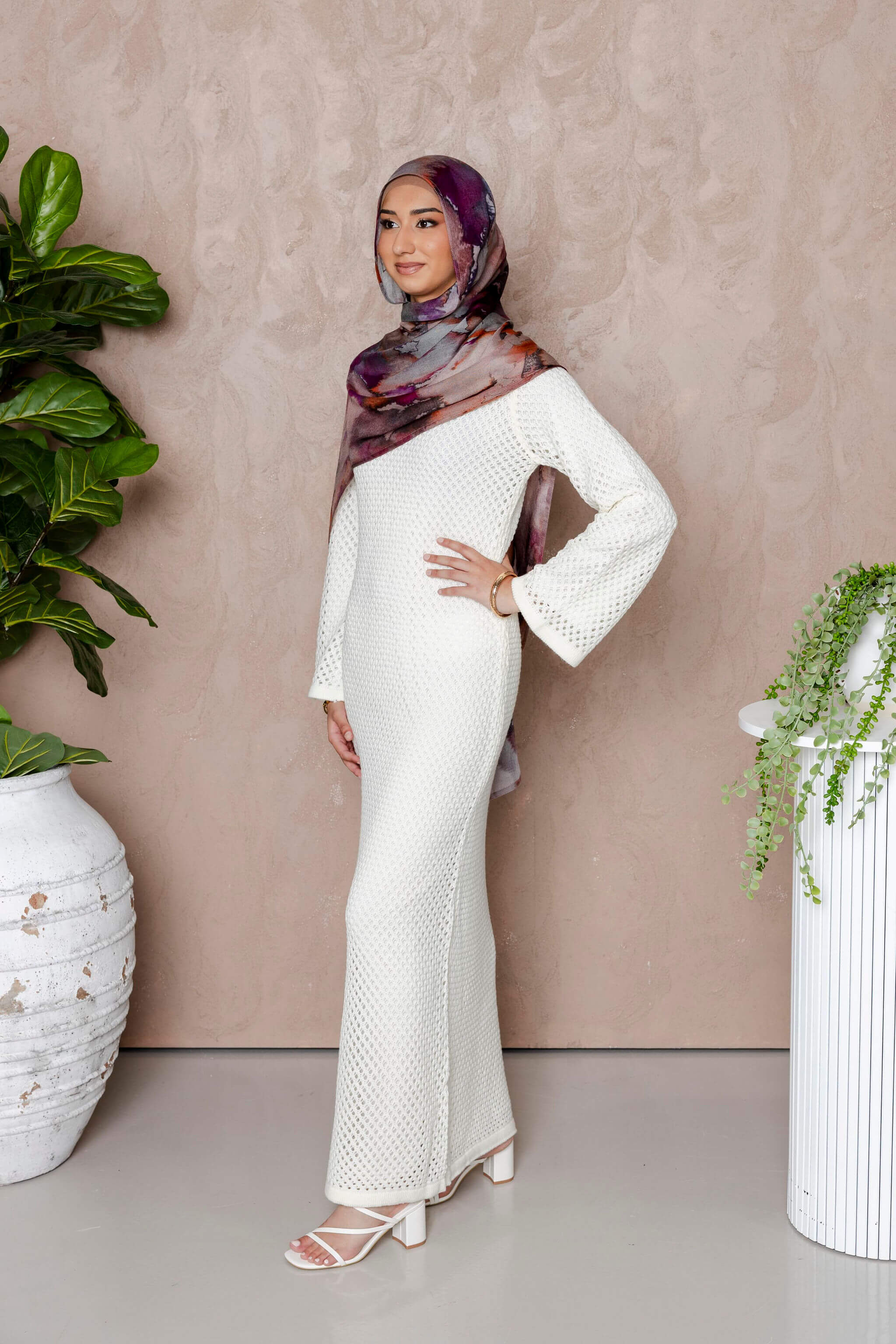 A woman in the Modest Goss Noor Knit Maxi Dress (White) and a patterned hijab stands sideways, smiling between two large potted plants against a neutral beige background.