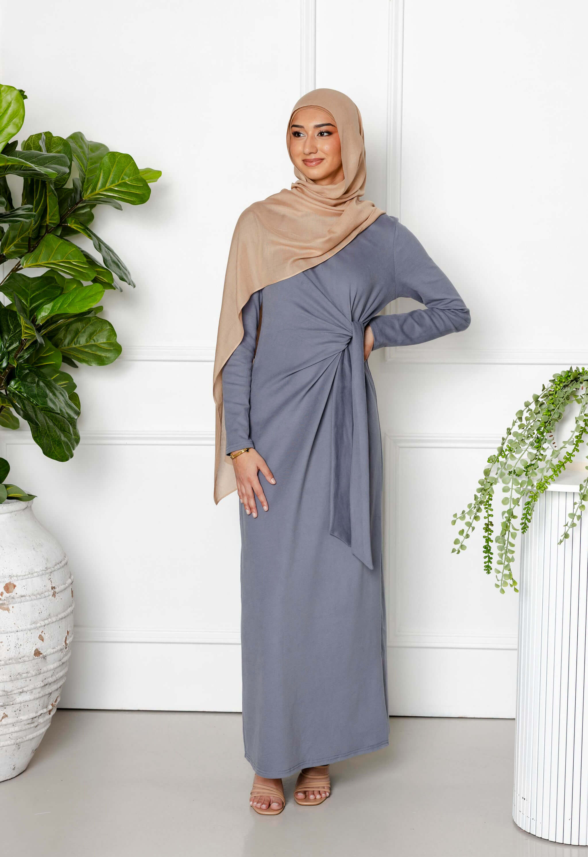 A woman in a taupe hijab and Modest Goss’s Maya Tie-up Maxi Dress stands indoors among green plants and white vases, smiling softly with one hand on her hip, showcasing elegant modest fashion.