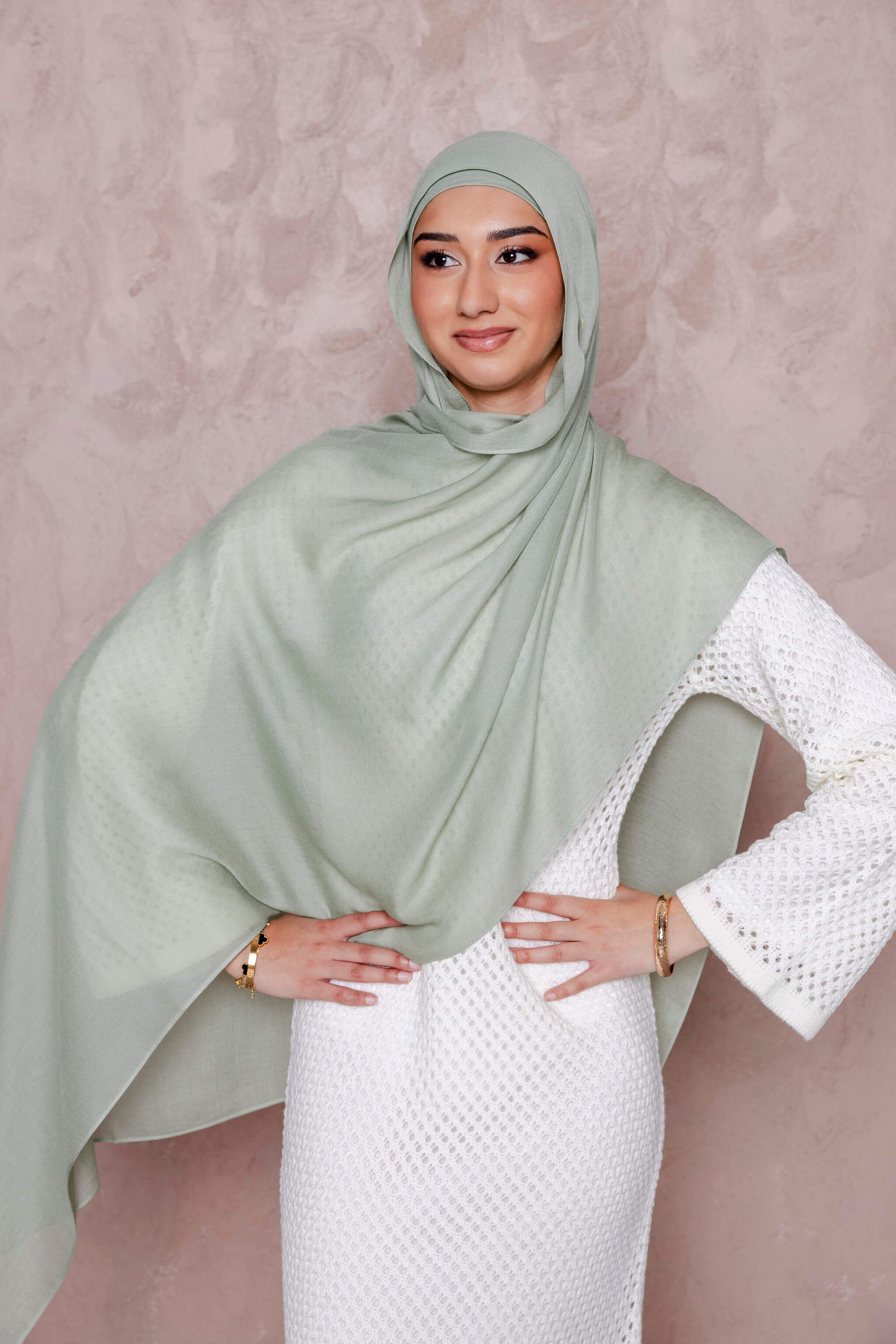 A woman in a Modest Goss #59 Smokey Blue lightweight, breathable hijab and a textured white dress stands against a pale pink background with her hands on her hips, smiling gently.