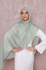 A woman in a Modest Goss #59 Smokey Blue lightweight, breathable hijab and a textured white dress stands against a pale pink background with her hands on her hips, smiling gently.