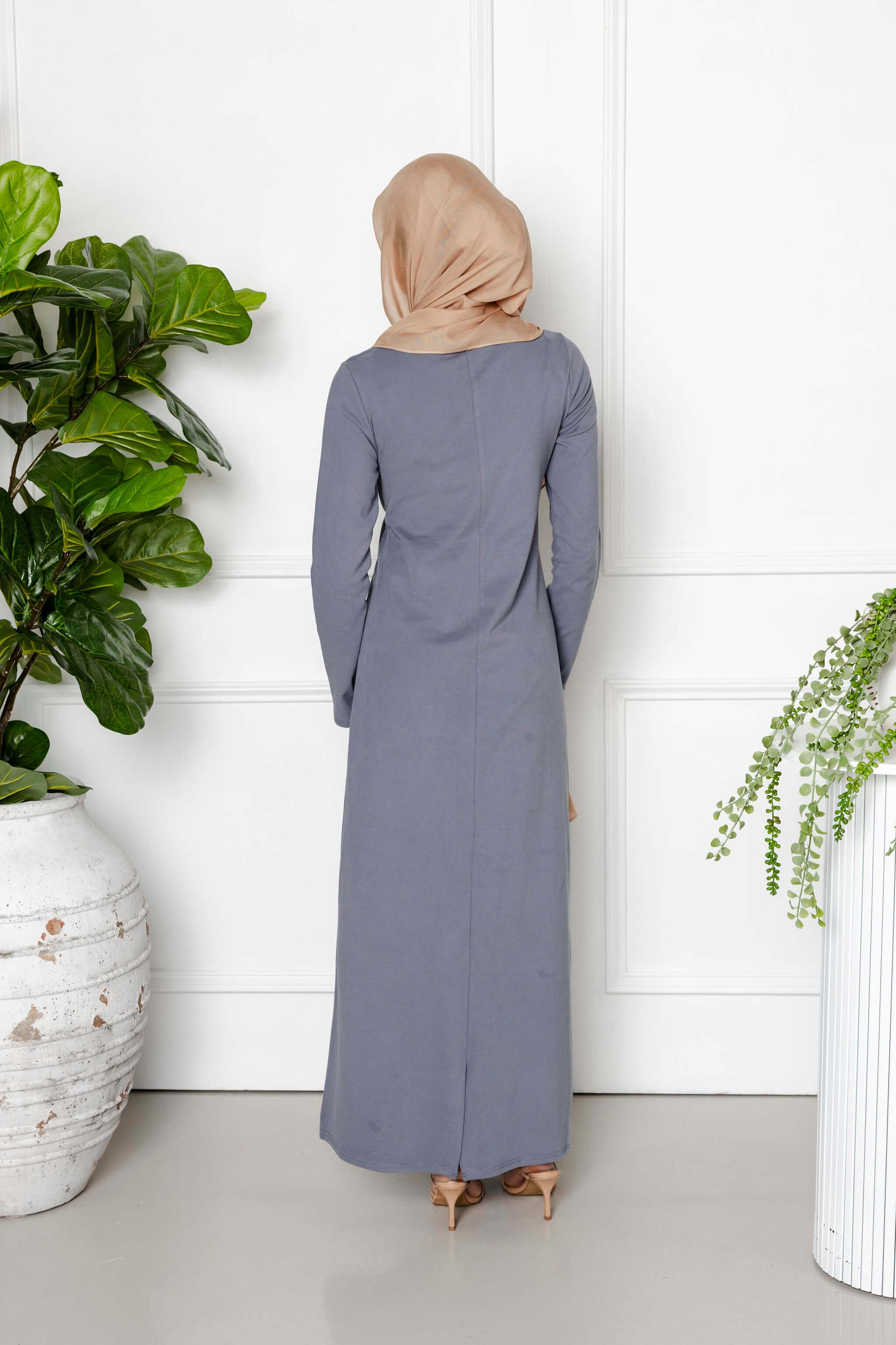 A woman in a light brown hijab and the Maya Tie-up Maxi Dress by Modest Goss faces away from the camera, surrounded by lush potted plants and a white wall, showcasing modern modest fashion.