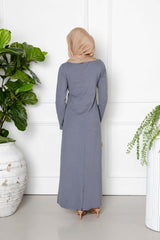 A woman in a light brown hijab and the Maya Tie-up Maxi Dress by Modest Goss faces away from the camera, surrounded by lush potted plants and a white wall, showcasing modern modest fashion.
