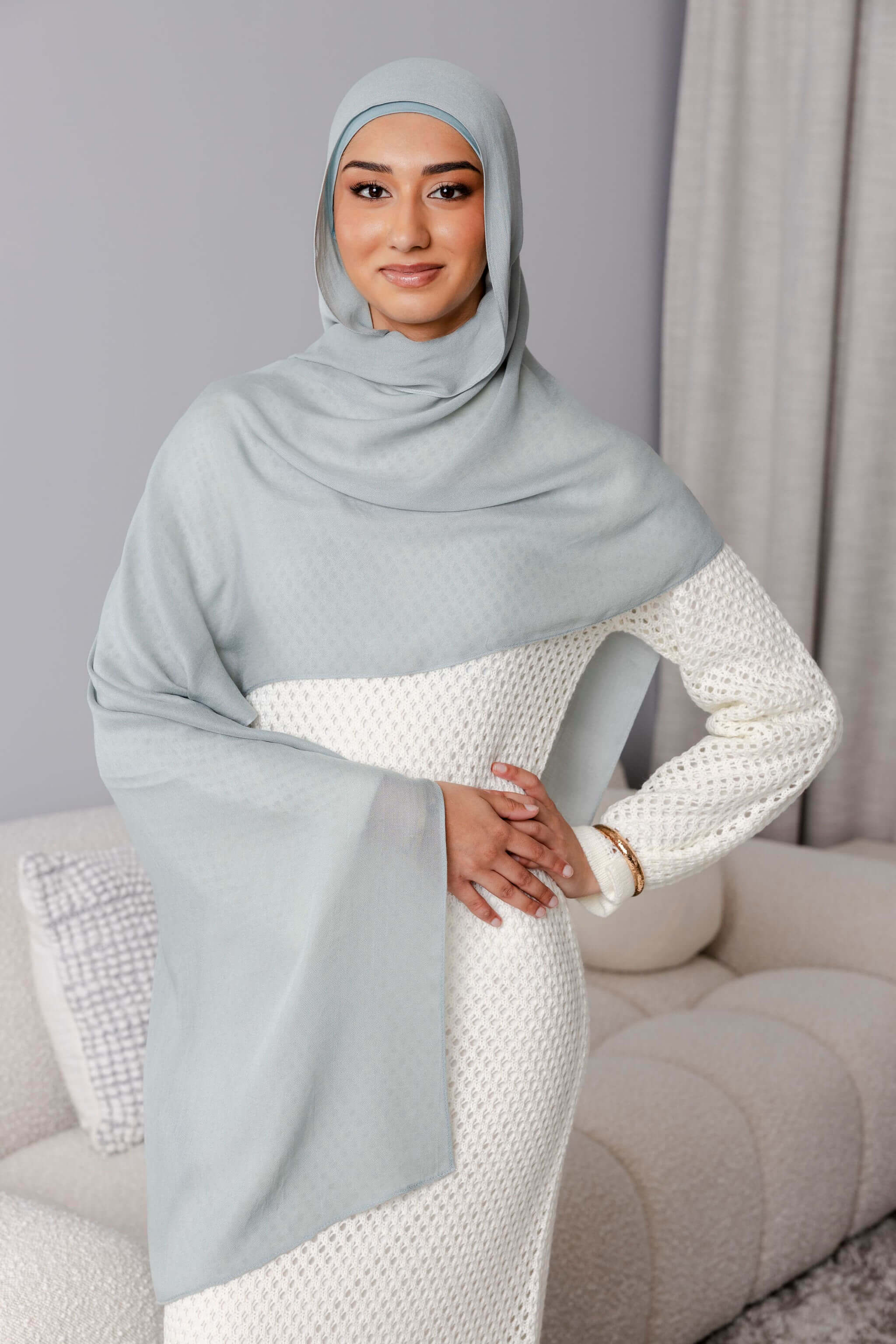 A woman wearing the Modest Goss #59 Smokey Blue Modal Hijab and a textured white dress stands with one hand on her hip, smiling softly in a modern, neutral-toned living room.