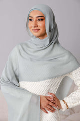 A woman models Modest Goss’s #59 Smokey Blue lightweight, breathable modal hijab with a matching under cap and a white textured dress, smiling softly with one hand on her hip against a plain background.