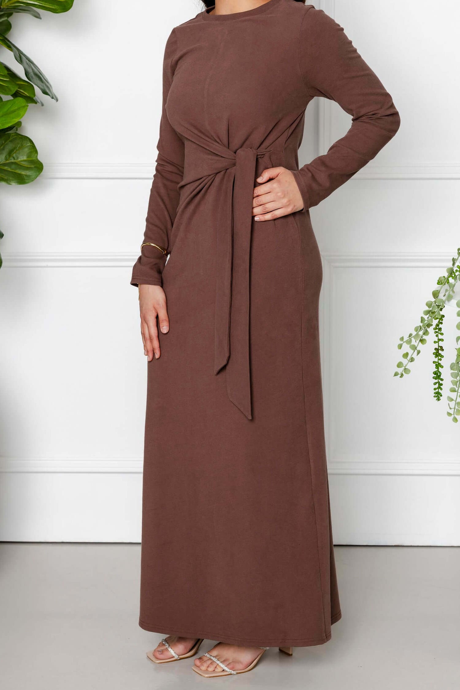 A woman poses indoors in the Modest Goss Maya Tie-up Maxi Dress, an ankle-length cocoa brown, long-sleeve dress with a front tie detail, paired with sandals beside potted green plants against a white wall.