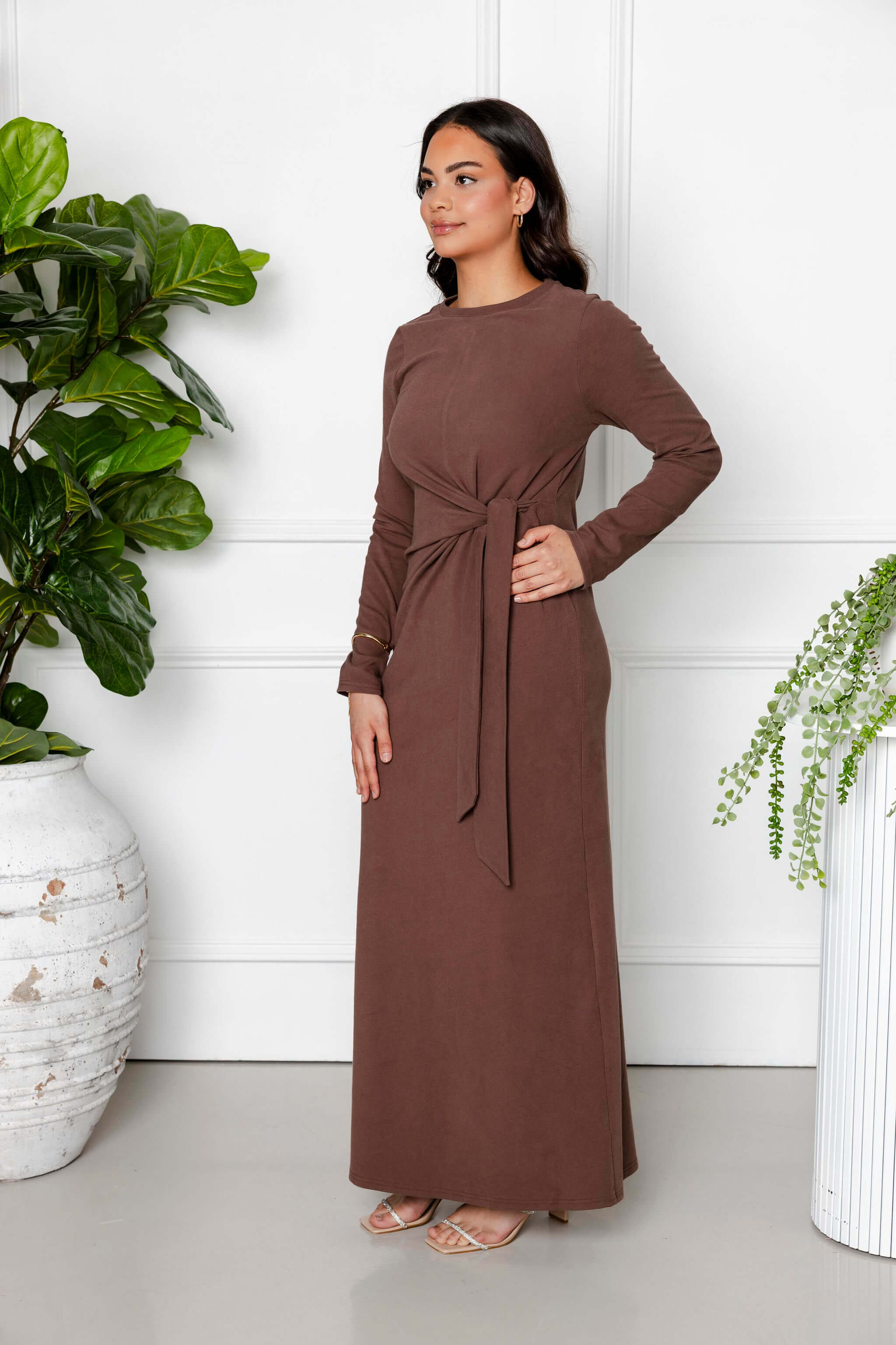 A woman poses indoors in the Modest Goss Maya Tie-up Maxi Dress, an ankle-length cocoa brown, long-sleeve dress with a front tie detail, paired with sandals beside potted green plants against a white wall.