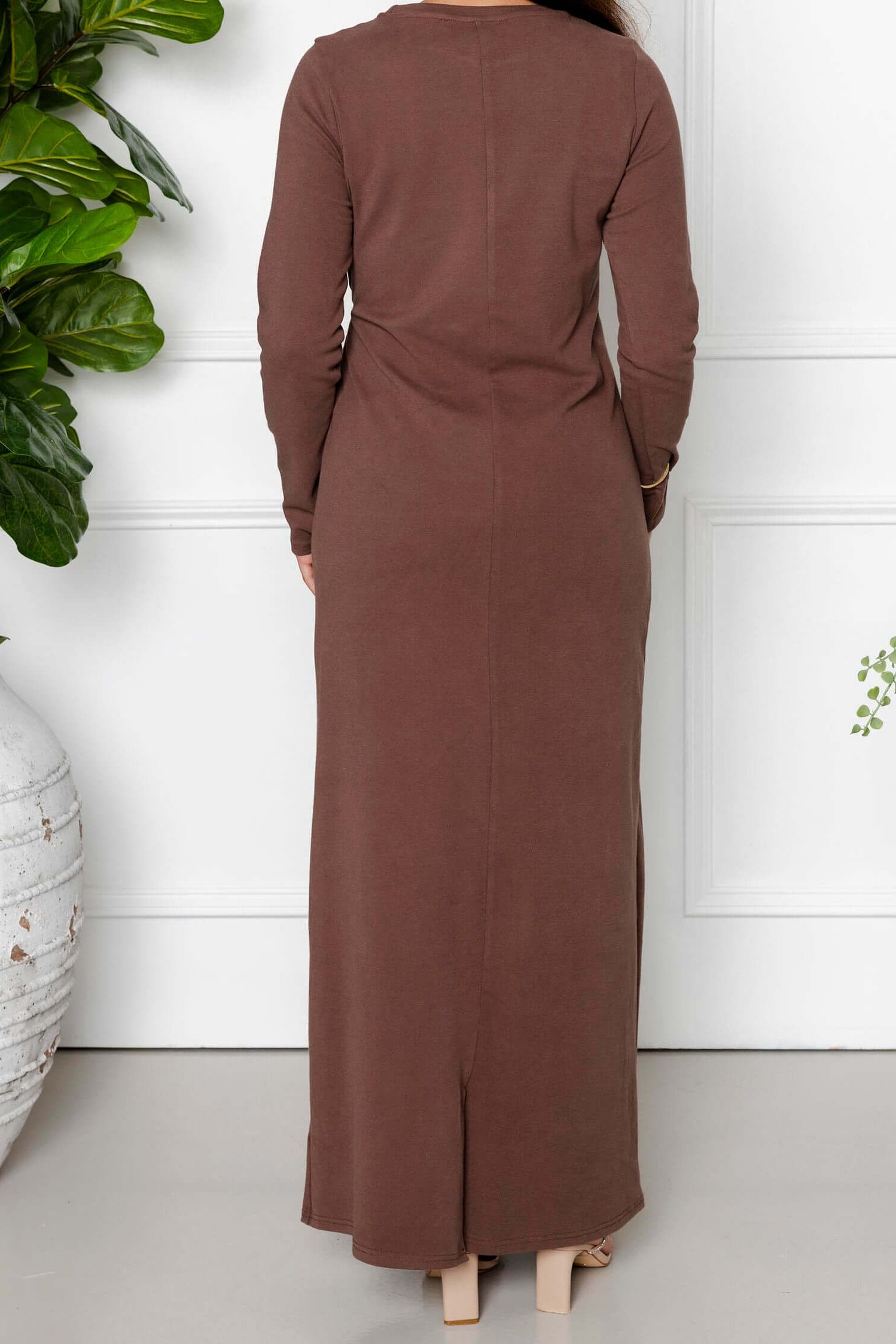 A woman with long dark hair faces away, wearing the Modest Goss Maya Tie-up Maxi Dress in cocoa brown. Indoors, she stands between two large potted plants against a white wall, showcasing modest fashion.