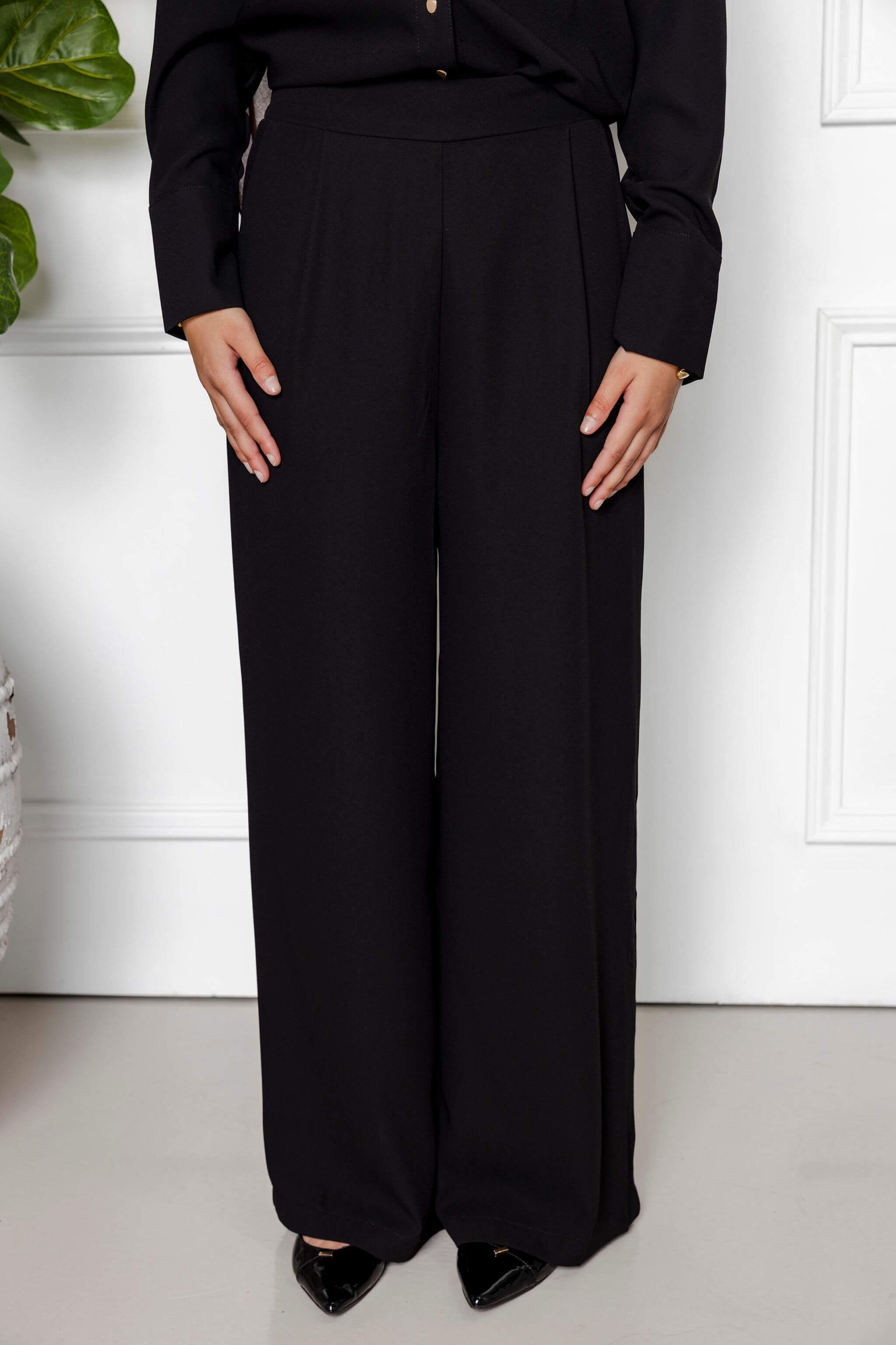 A person stands indoors wearing the Modest Goss Jasmine 2 Piece Set (Black), featuring wide-leg trousers and a long-sleeve top, paired with black pointed-toe shoes for a sleek, modest look.