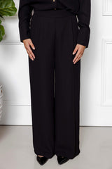 A person stands indoors wearing the Modest Goss Jasmine 2 Piece Set (Black), featuring wide-leg trousers and a long-sleeve top, paired with black pointed-toe shoes for a sleek, modest look.