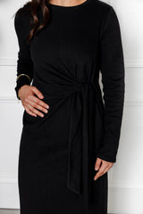 A woman models the Modest Goss Maya Tie-up Maxi Dress—a long-sleeved black dress with a waist knot—standing against a white background, her left hand on her hip and dark hair over one shoulder, showcasing contemporary modest style.