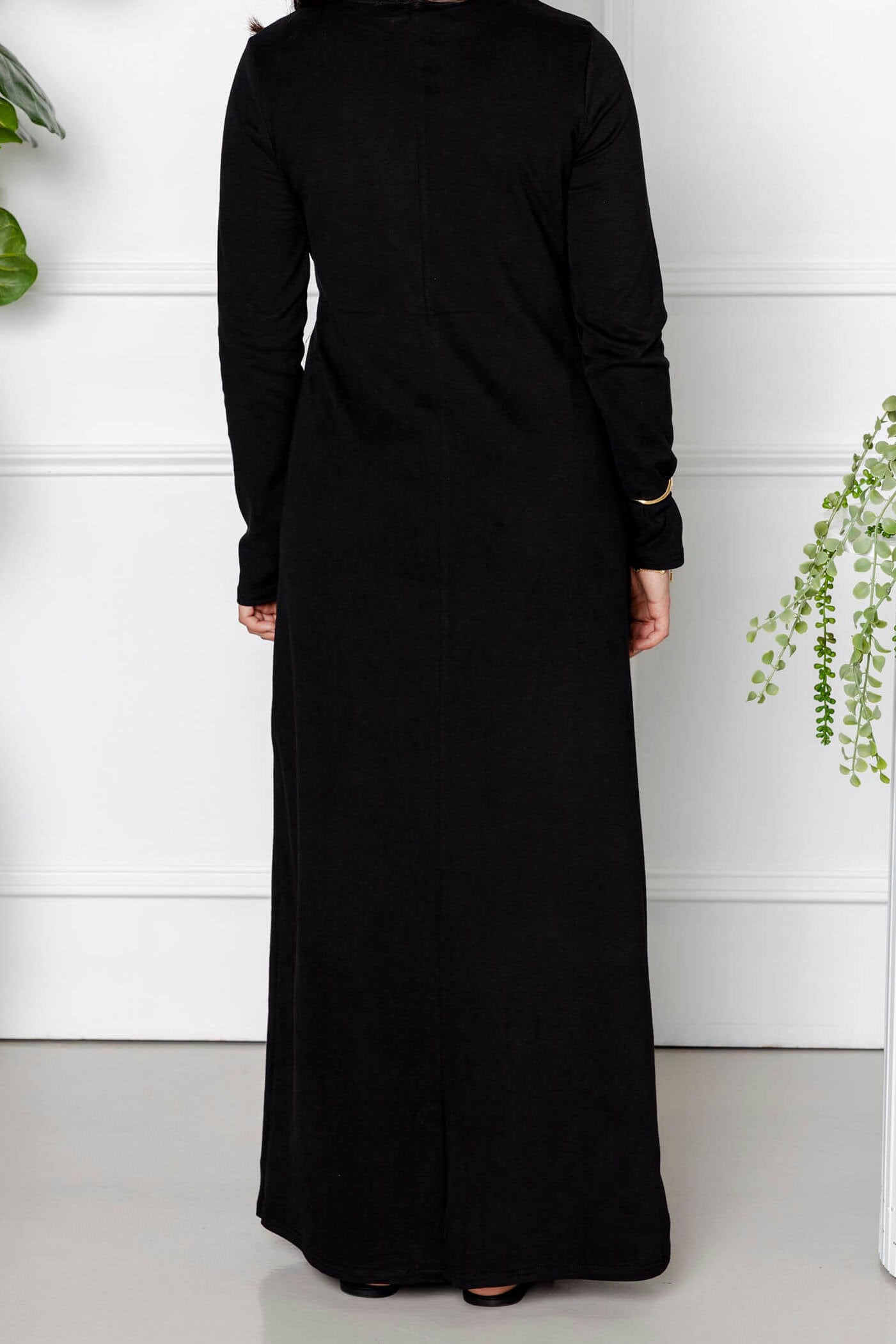 A woman stands indoors with her back to the camera, wearing the Modest Goss Maya Tie-up Maxi Dress with long sleeves. She is between two large potted plants, set against a white wall with decorative paneling.