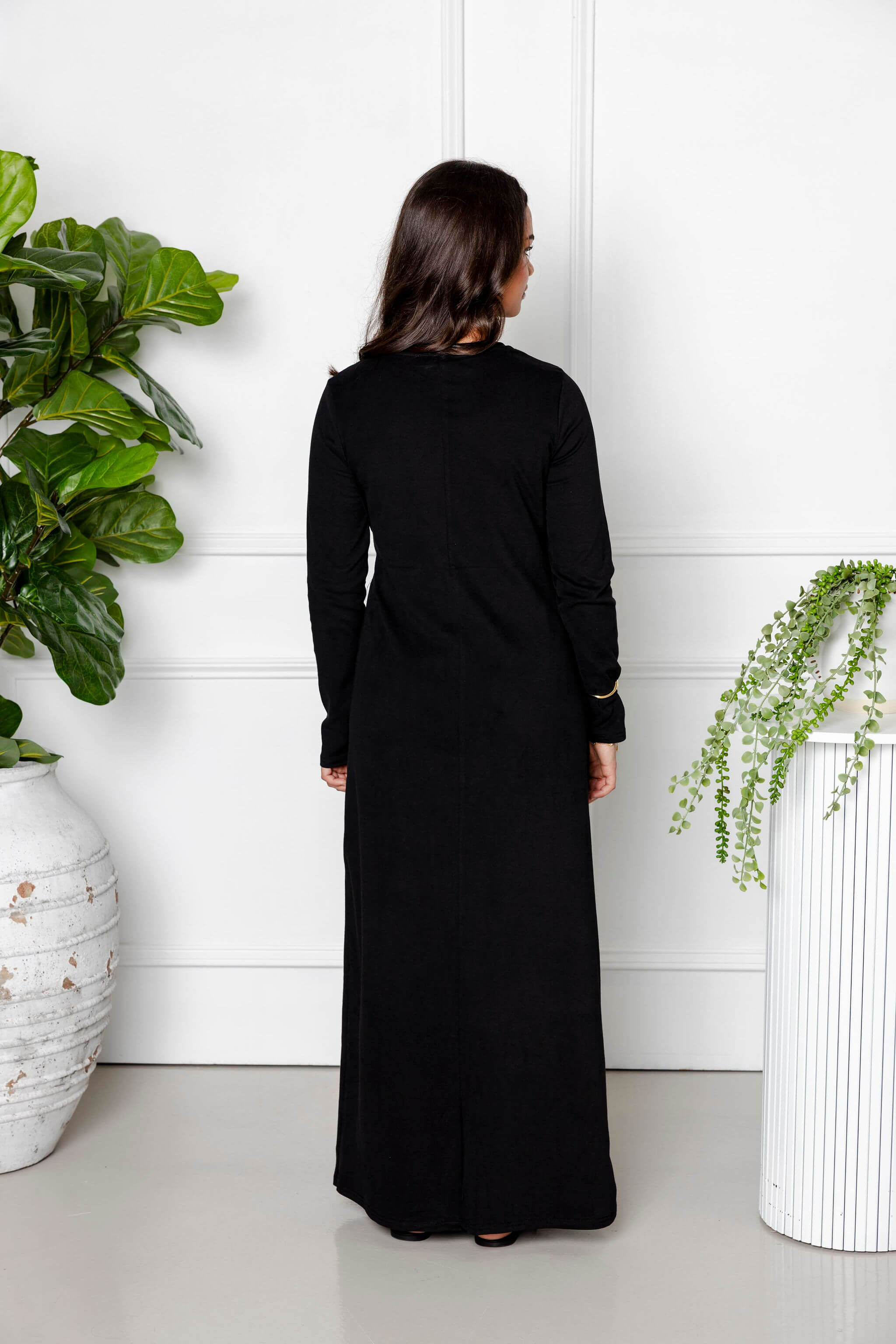 A woman stands indoors with her back to the camera, wearing the Modest Goss Maya Tie-up Maxi Dress with long sleeves. She is between two large potted plants, set against a white wall with decorative paneling.