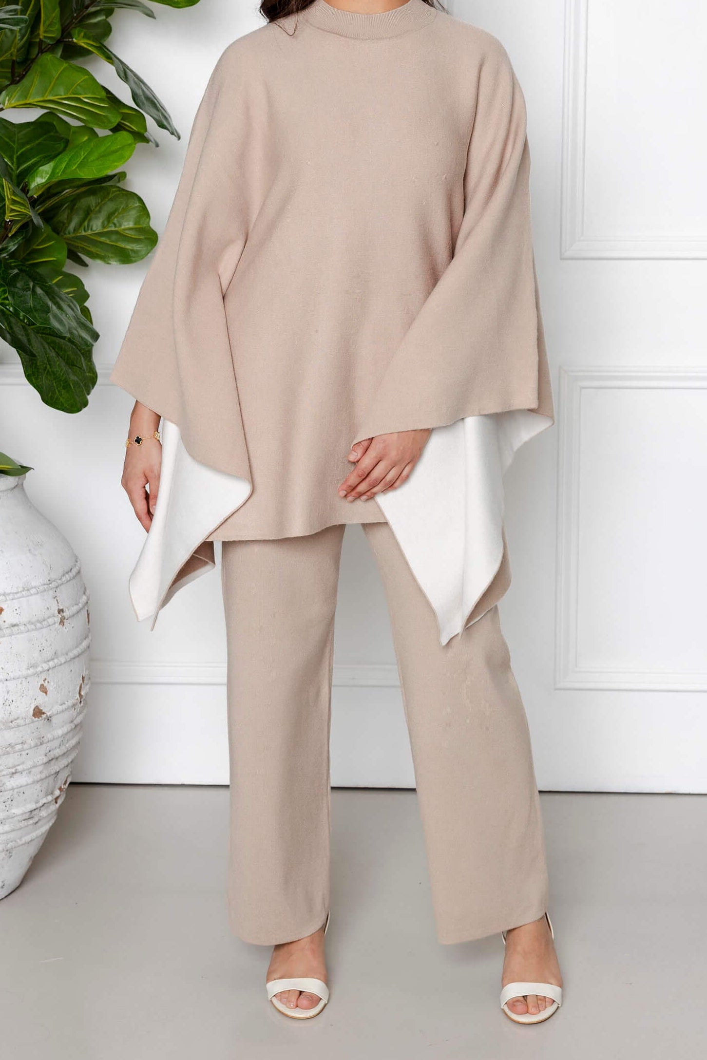 A woman stands indoors among potted plants, exuding calmness as she wears the Modest Goss Amara Knit Poncho 2 Piece Set in beige, featuring a modest knit poncho with white lining and matching pants.