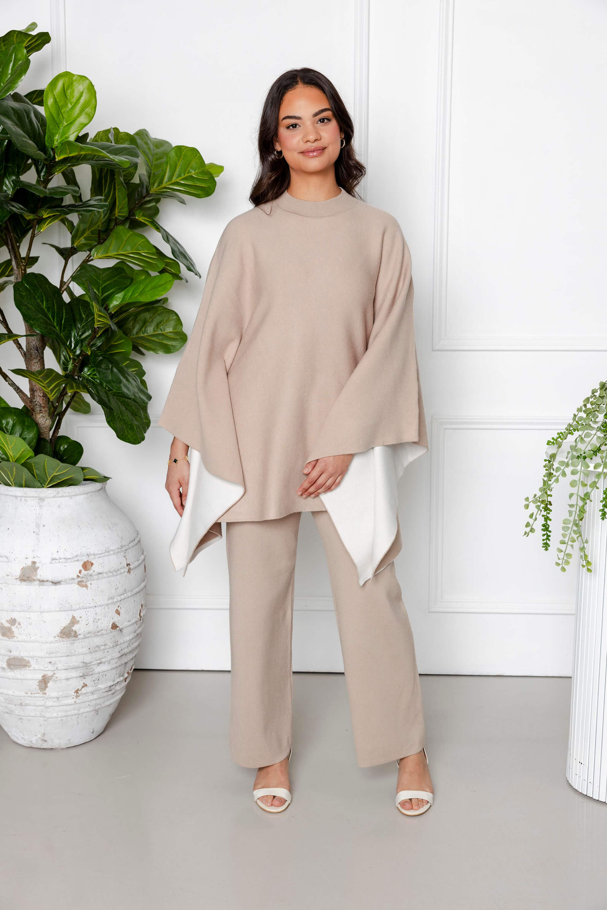 A woman stands indoors among potted plants, exuding calmness as she wears the Modest Goss Amara Knit Poncho 2 Piece Set in beige, featuring a modest knit poncho with white lining and matching pants.