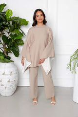 A woman stands indoors among potted plants, exuding calmness as she wears the Modest Goss Amara Knit Poncho 2 Piece Set in beige, featuring a modest knit poncho with white lining and matching pants.