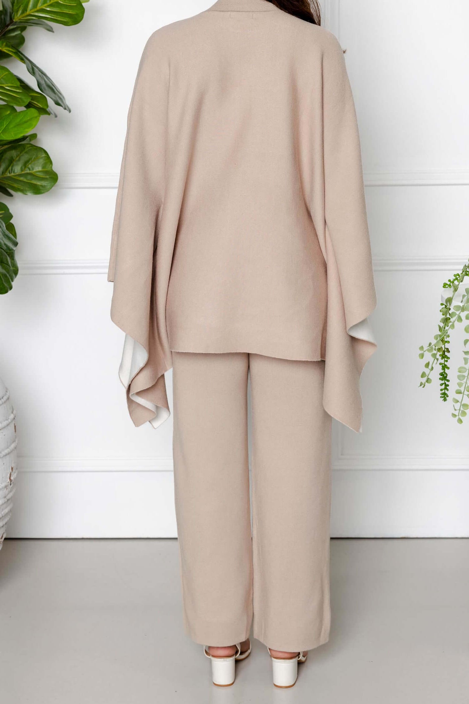 A woman with long dark hair stands indoors, facing away, wearing the Modest Goss Amara Knit Poncho 2 Piece Set in beige, featuring wide sleeves and pants. She is surrounded by green potted plants against a white wall.