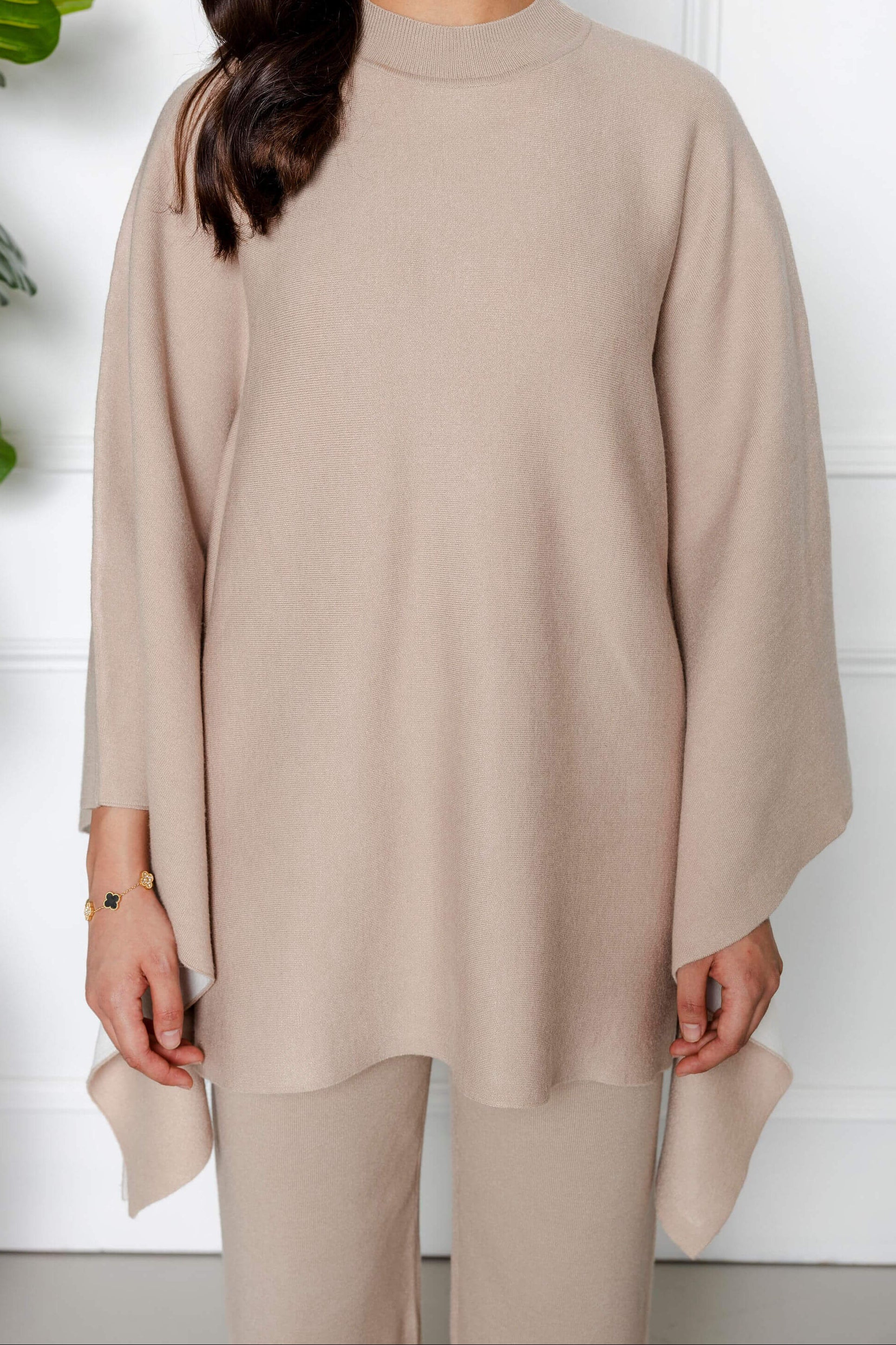 A woman models the Modest Goss Amara Knit Poncho 2 Piece Set in beige—featuring a loose, wide-sleeved top and matching pants. She stands indoors against a white background; her hair is partly visible while her face is not shown.