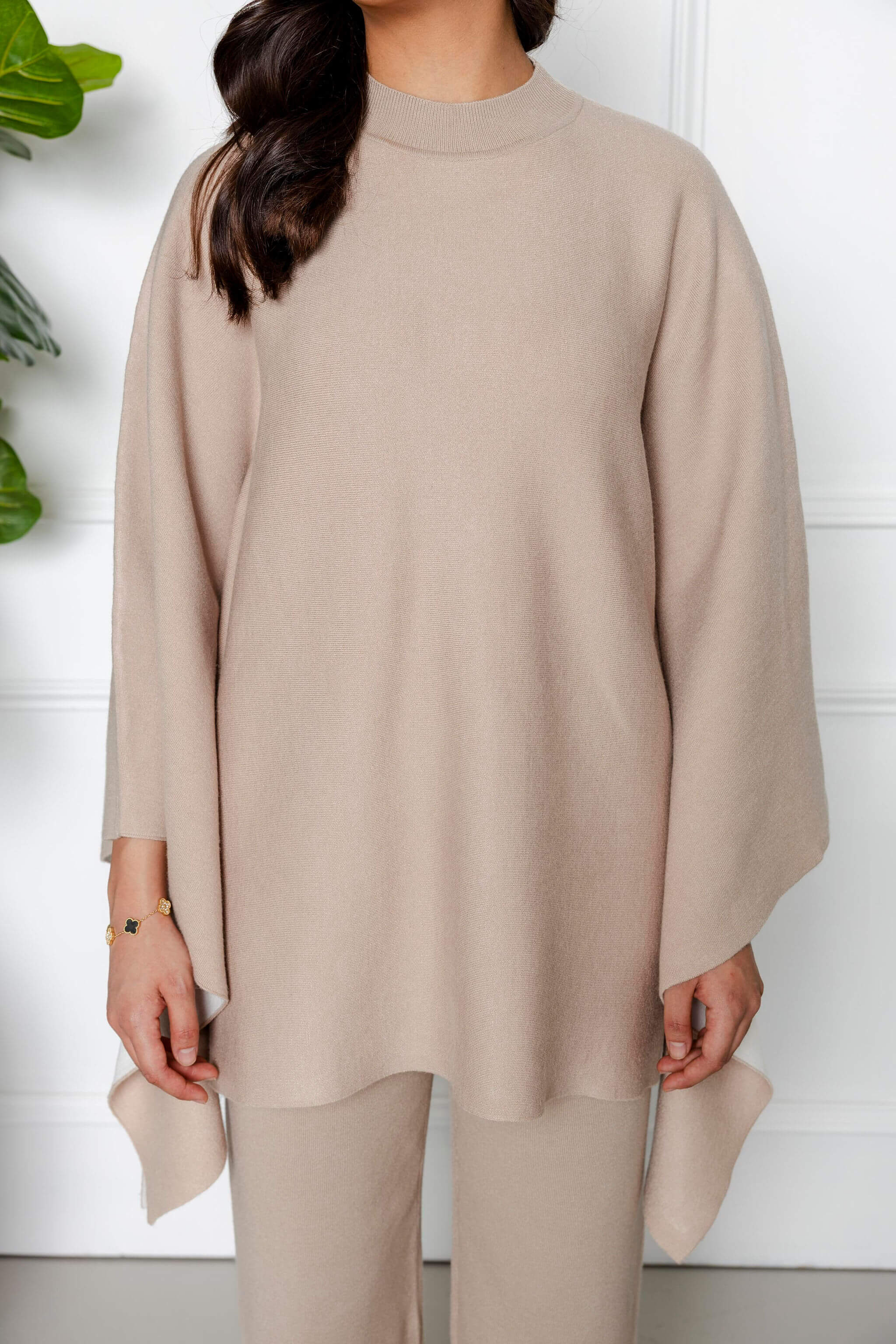 A woman models the Modest Goss Amara Knit Poncho 2 Piece Set in beige—featuring a loose, wide-sleeved top and matching pants. She stands indoors against a white background; her hair is partly visible while her face is not shown.
