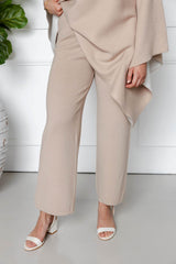 A person wearing the Modest Goss Amara Knit Poncho 2 Piece Set in beige and white open-toed sandals stands indoors against a white wall, with only their lower half visible.