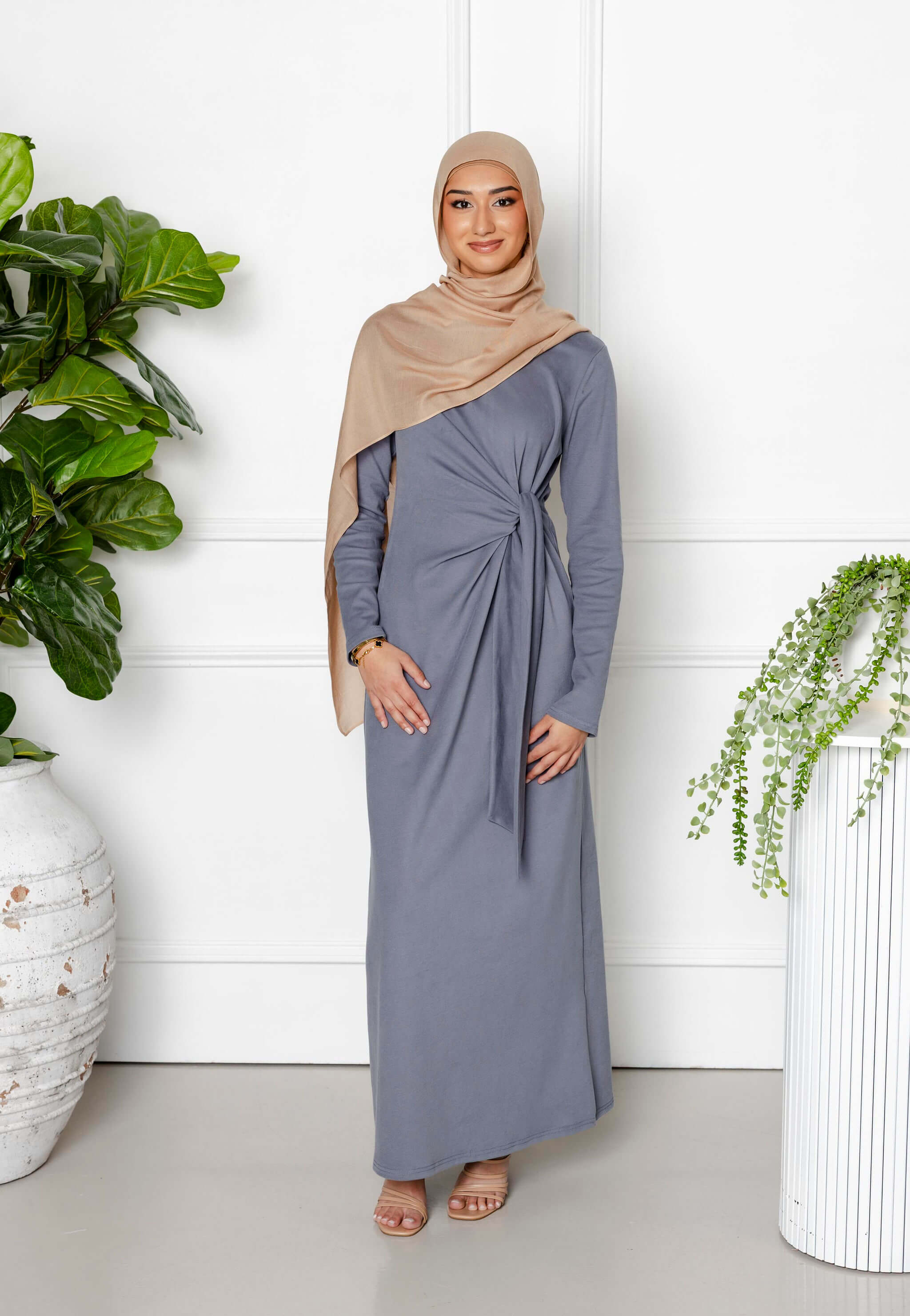 A woman in a beige hijab and the Maya Tie-up Maxi Dress by Modest Goss stands indoors between two large potted plants, smiling at the camera against a white wall, showcasing elegant modest fashion.
