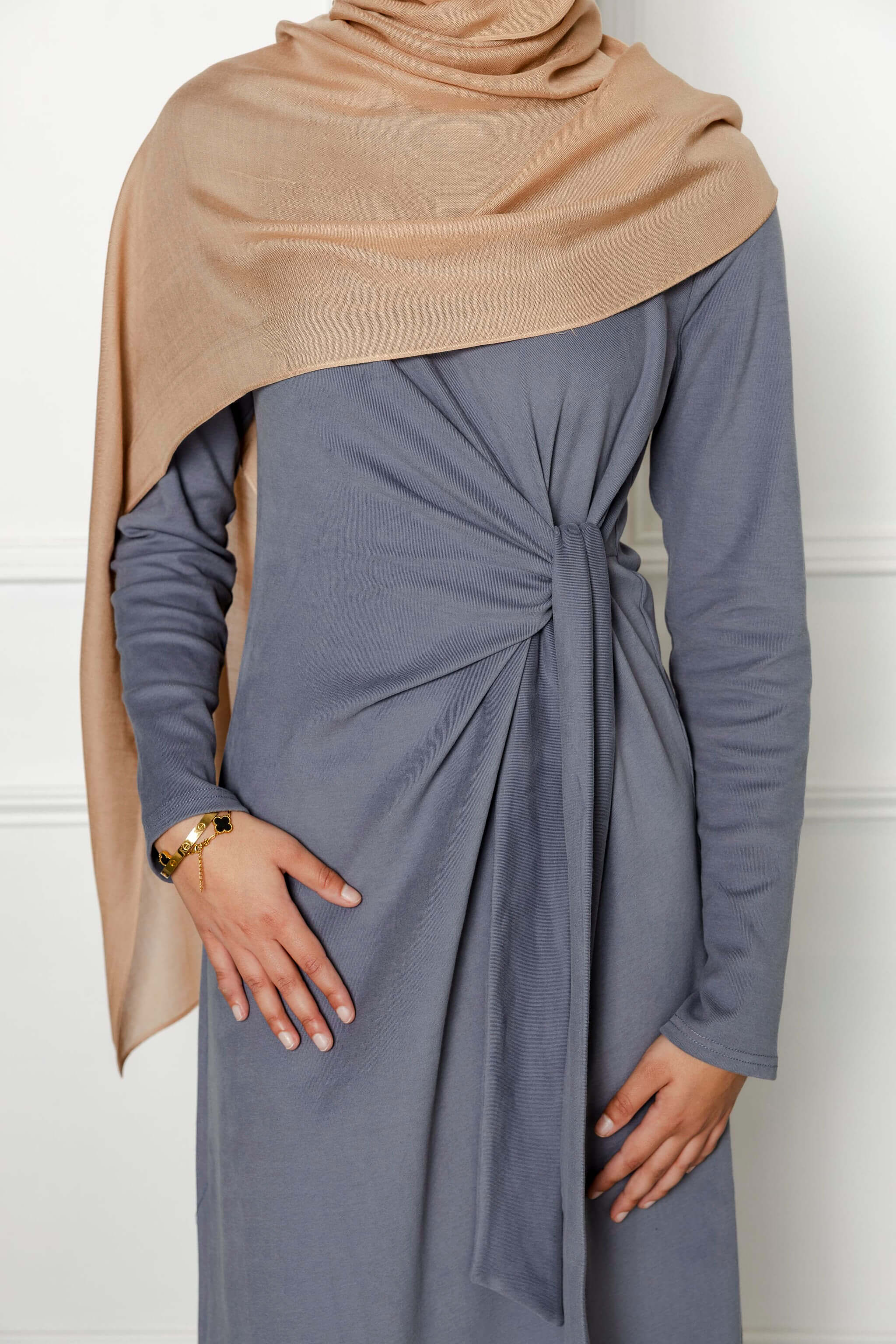 A person wears the Modest Goss Maya Tie-up Maxi Dress in gray with a front knot and a light brown hijab; left hand on hip displaying a gold watch, right hand by their side. The white background showcases modest fashion style.