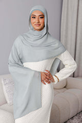 A woman wearing the Modest Goss #59 Smokey Blue Modal Hijab and a textured white dress stands with one hand on her hip, smiling softly in a modern, neutral-toned living room.