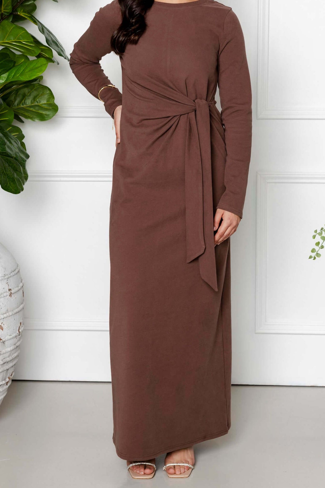 A woman stands indoors before white paneled walls, smiling softly in the Maya Tie-up Maxi Dress by Modest Goss in cocoa brown. Her long, wavy dark hair flows as she embraces modest fashion amidst large potted plants.