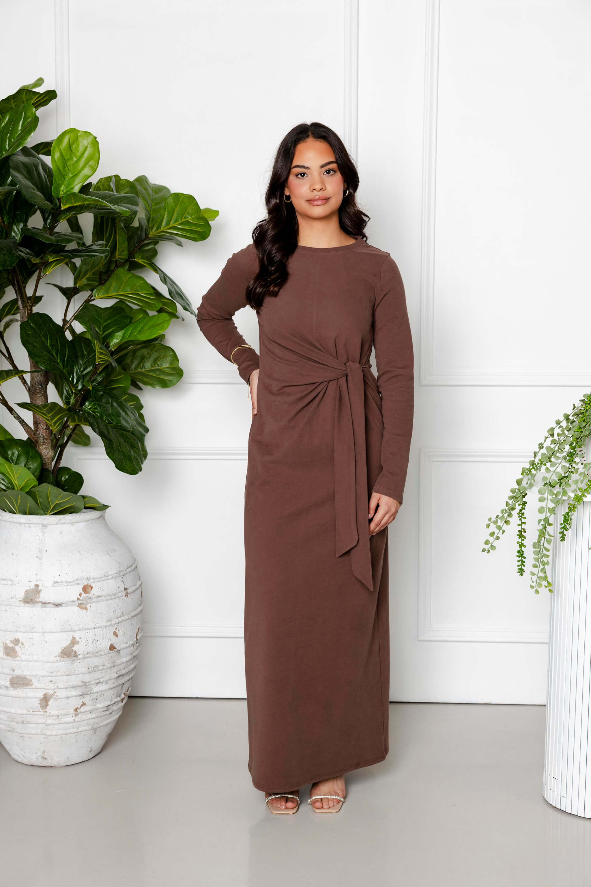 A woman stands indoors before white paneled walls, smiling softly in the Maya Tie-up Maxi Dress by Modest Goss in cocoa brown. Her long, wavy dark hair flows as she embraces modest fashion amidst large potted plants.
