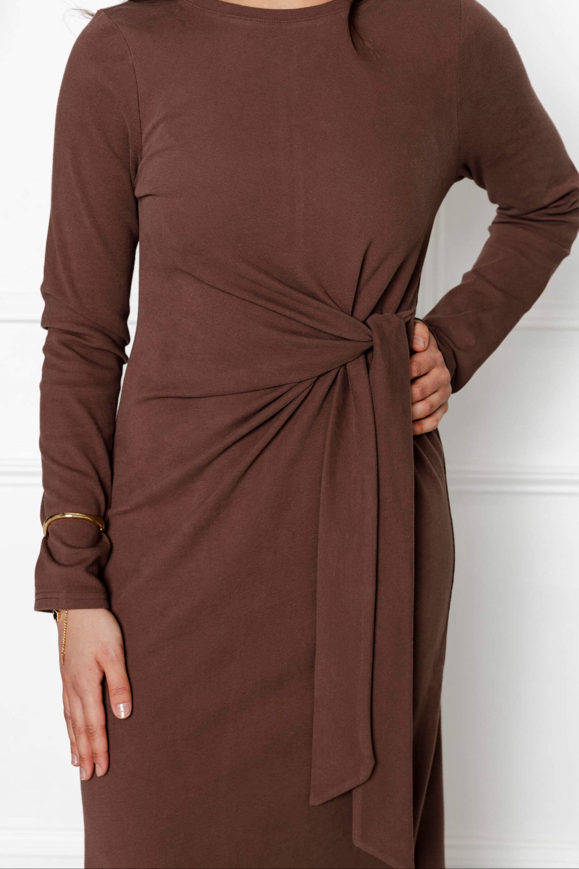 Wearing the Modest Goss Maya Tie-up Maxi Dress—a cocoa brown, long-sleeved maxi with a waist knot—this person poses with their right hand on hip and a gold bracelet. Face not visible; a chic example of modest fashion.