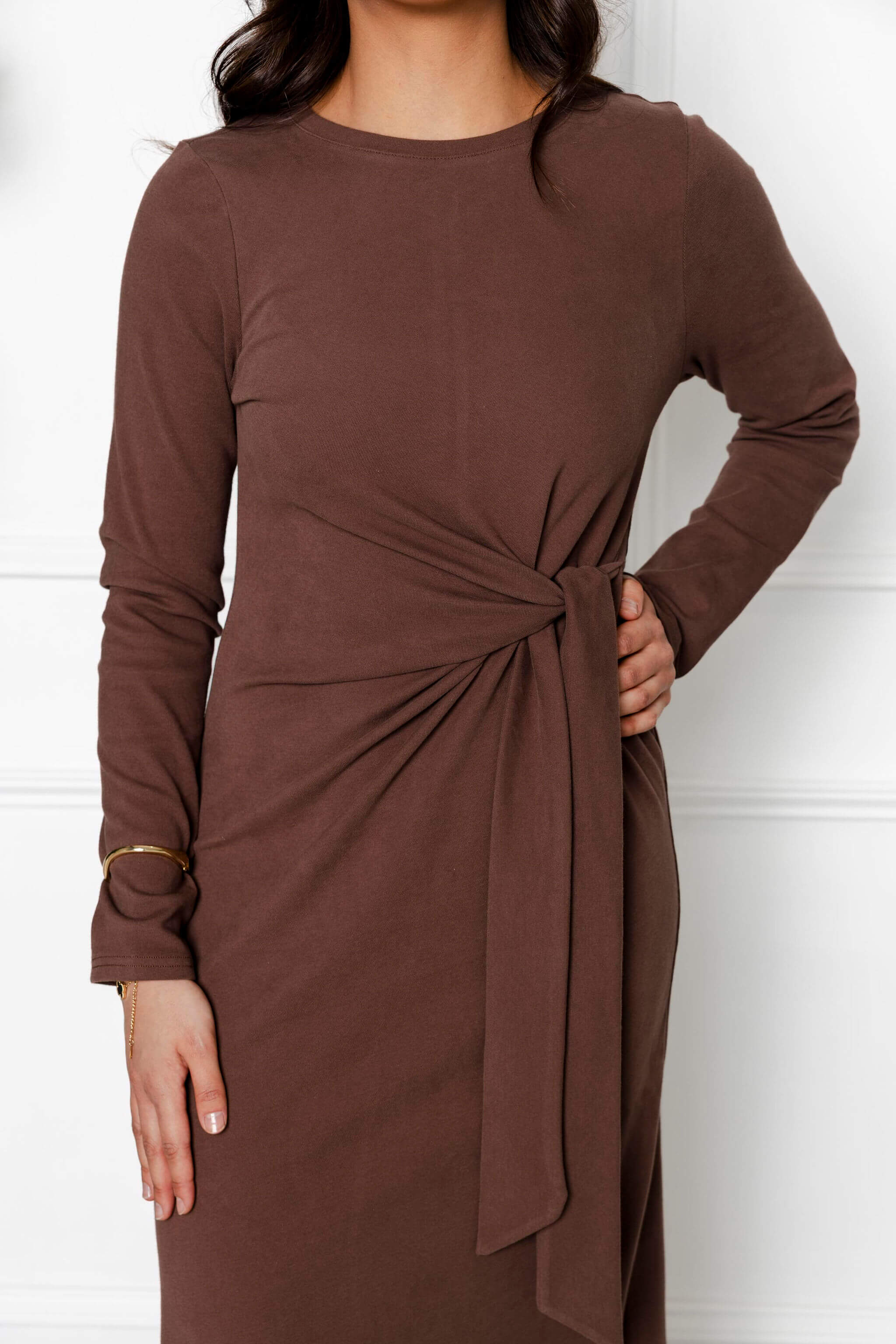 Wearing the Modest Goss Maya Tie-up Maxi Dress—a cocoa brown, long-sleeved maxi with a waist knot—this person poses with their right hand on hip and a gold bracelet. Face not visible; a chic example of modest fashion.