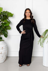 A woman in the Maya Tie-up Maxi Dress by Modest Goss stands indoors between large potted plants, one hand on a planter. Her look features modest fashion with long sleeves, dark hair, and black sandals.